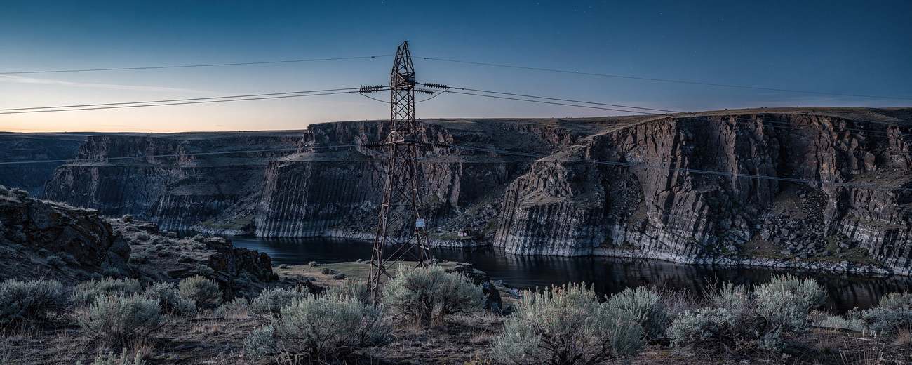 A transmission tower crossing a basalt river canyon at deep dusk.